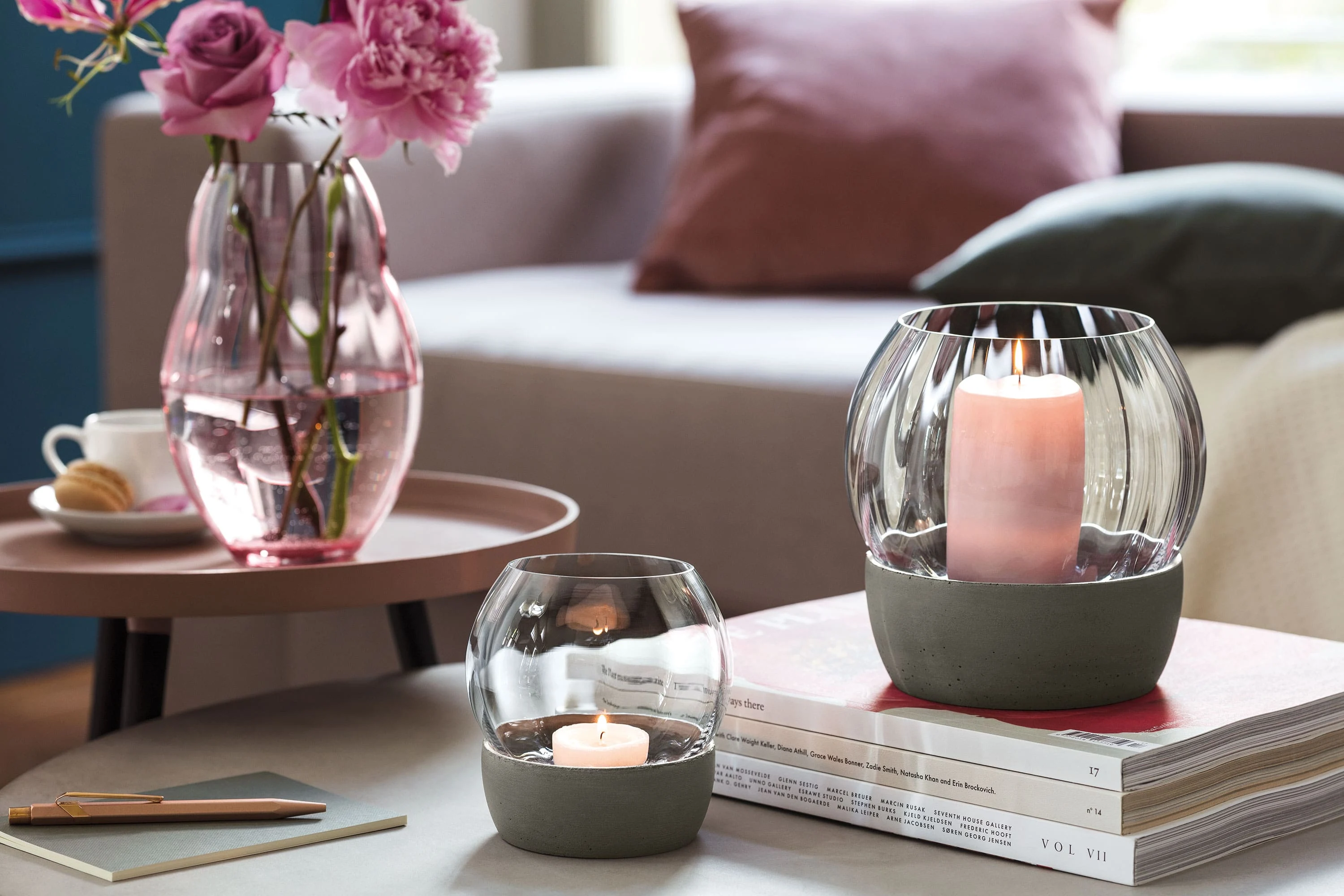 Zwei Rose Garden Windlichter von Villeroy & Boch stehen auf einem Stapel Bücher neben einer rosa Rose Garden Vase mit Blumen, alles auf einem runden Tisch in einem gemütlichen Wohnzimmerambiente.	