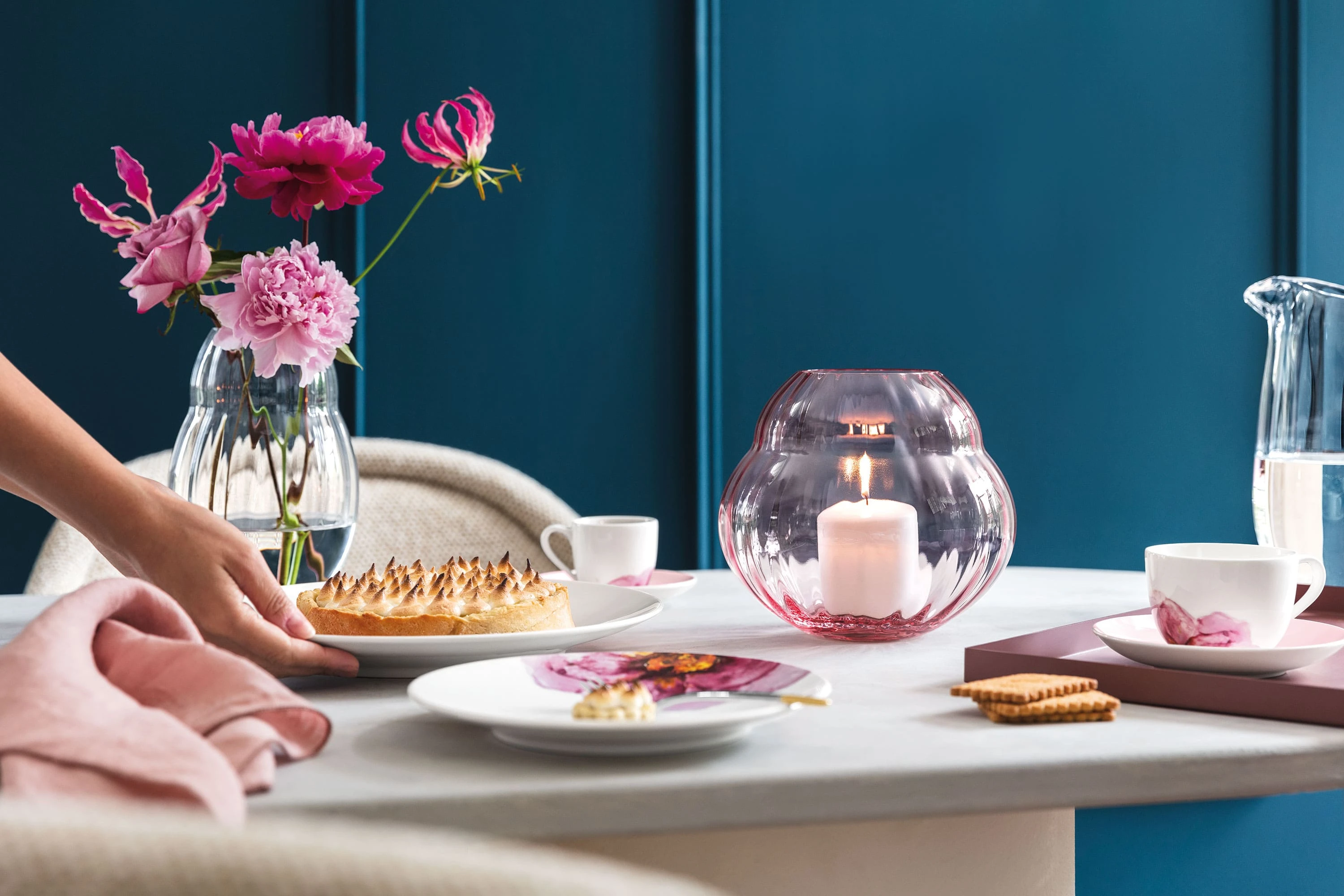 Une table de salle à manger élégante avec une personne servant le dessert, des fleurs fraîches dans des vases Rose Garden et des assiettes de la collection Rose Garden qui accentuent l’ambiance.