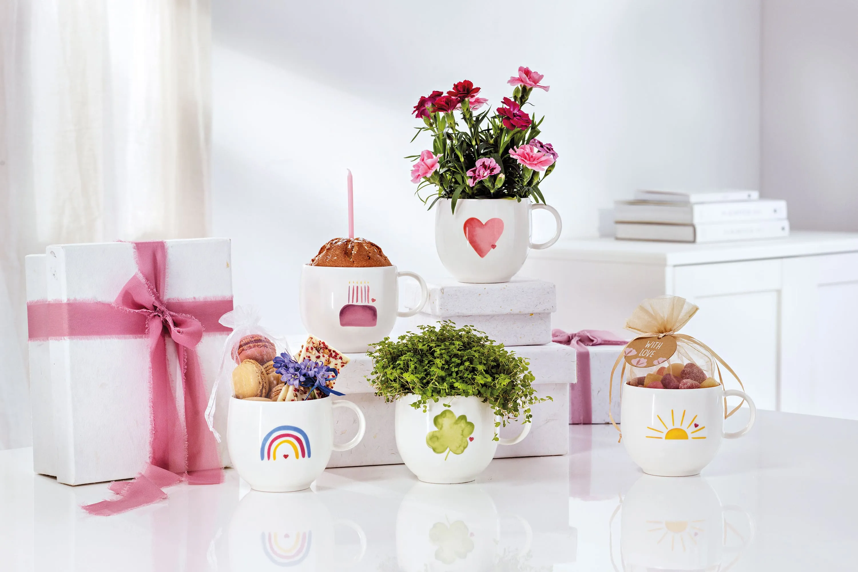 Cinq tasses blanches « With Love » de Villeroy & Boch, chacune avec un motif différent de fleurs, de sucreries et de cadeaux, sont posées sur une surface blanche. En arrière-plan, deux cadeaux emballés avec des rubans roses complètent cette scène enchanteresse.