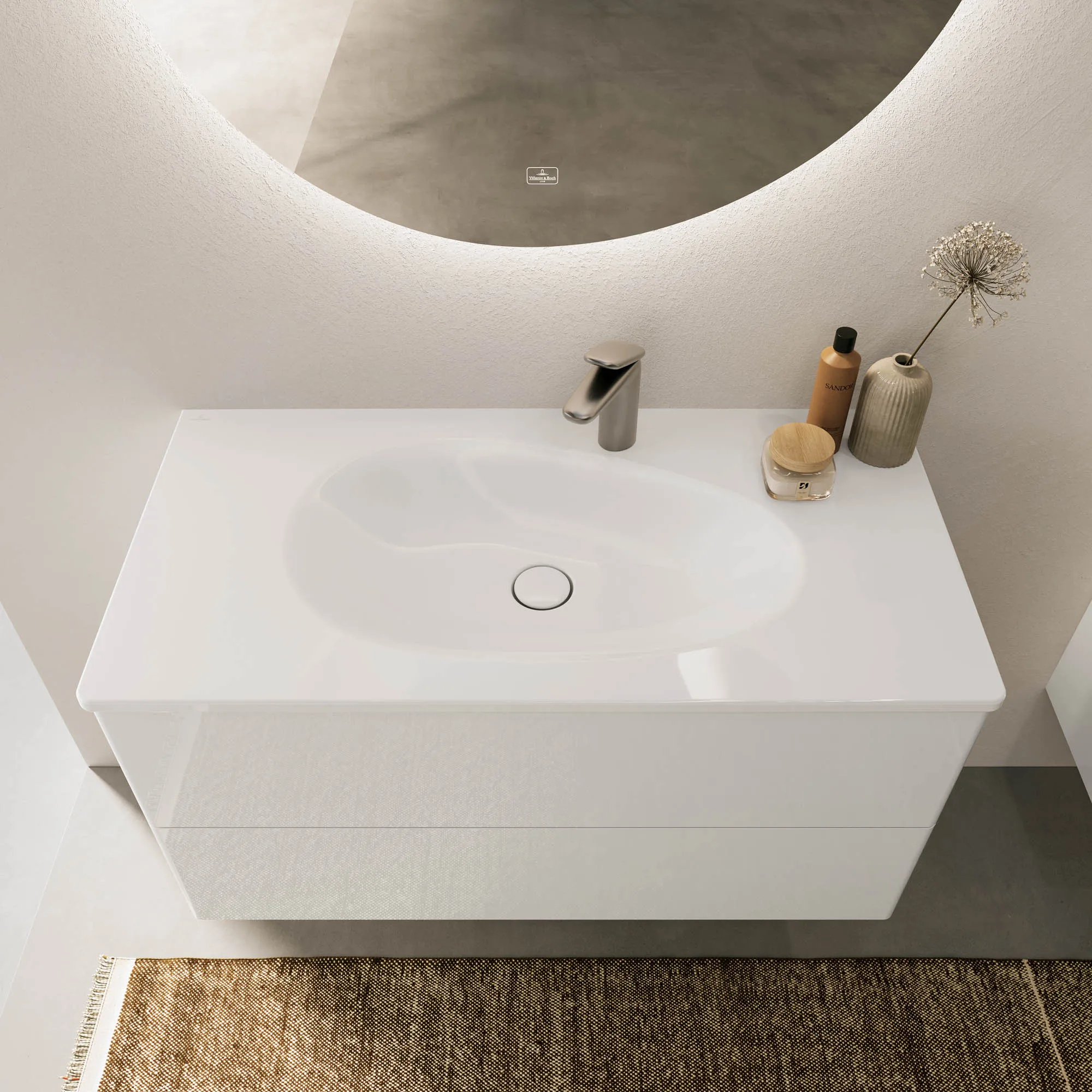 Un meuble sous plan avec un lavabo en blanc brillant ainsi qu’un miroir, de la décoration et un distributeur de savon Villeroy & Boch.