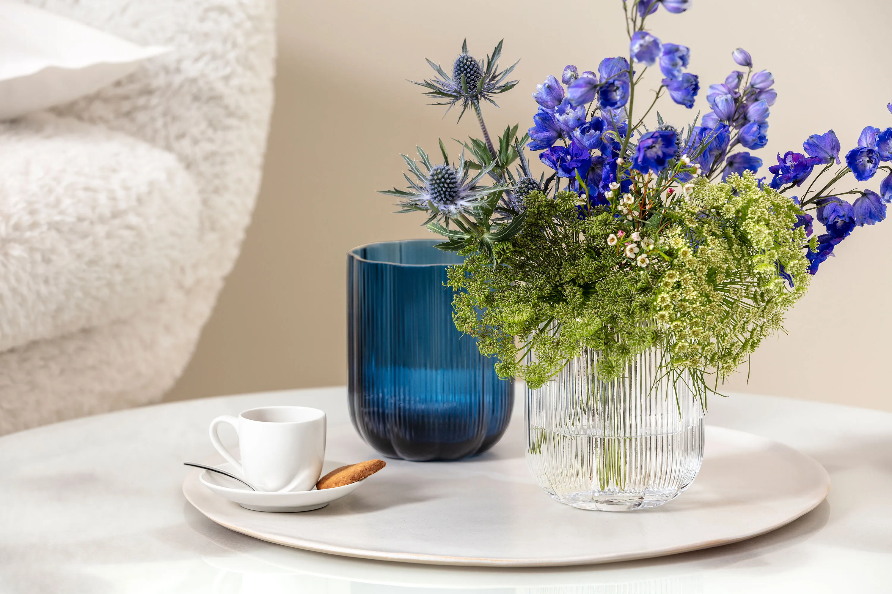 Eine kleine weiße For me Tasse mit einem Keks auf einer Untertasse, neben einer blauen Fleur Vase und einer durchsichtigen Fleur Vase Eine kleine weiße For me Tasse mit einem Keks auf einer Untertasse, neben einer blauen Fleur Vase und einer durchsichtigen Fleur Vase mit blauen und grünen Blumen auf einer runden Marmoroberfläche.