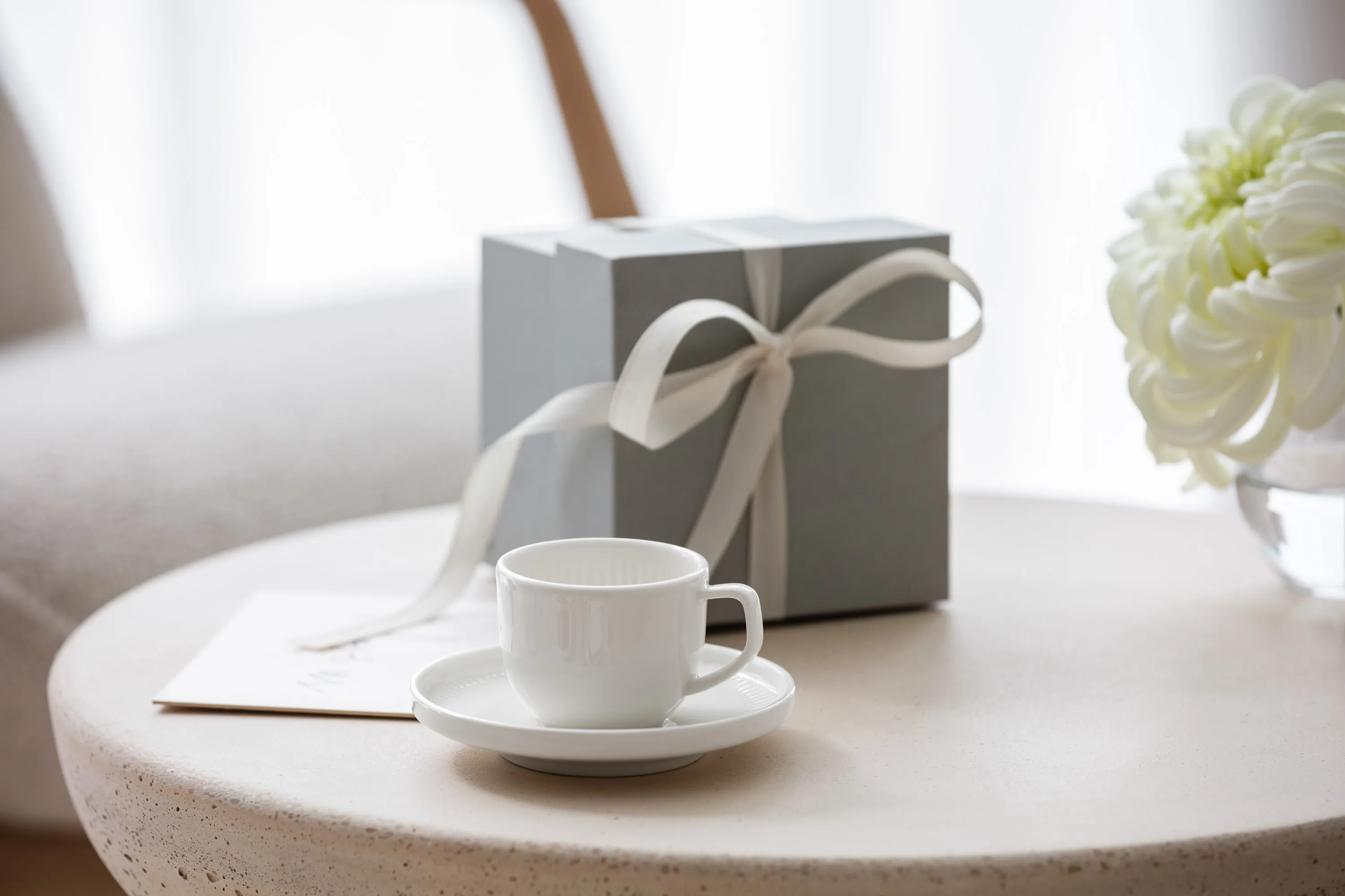 Une tasse Afina blanche sur une sous-tasse, une boîte cadeau avec un ruban et un bouquet de fleurs blanches sur une table ronde.