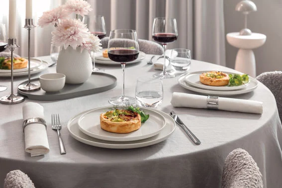 Ein Villeroy & Boch Afina-Tischset für vier Personen mit Quichetellern, Weingläsern, Servietten und einer Vase mit rosa Blumen.
