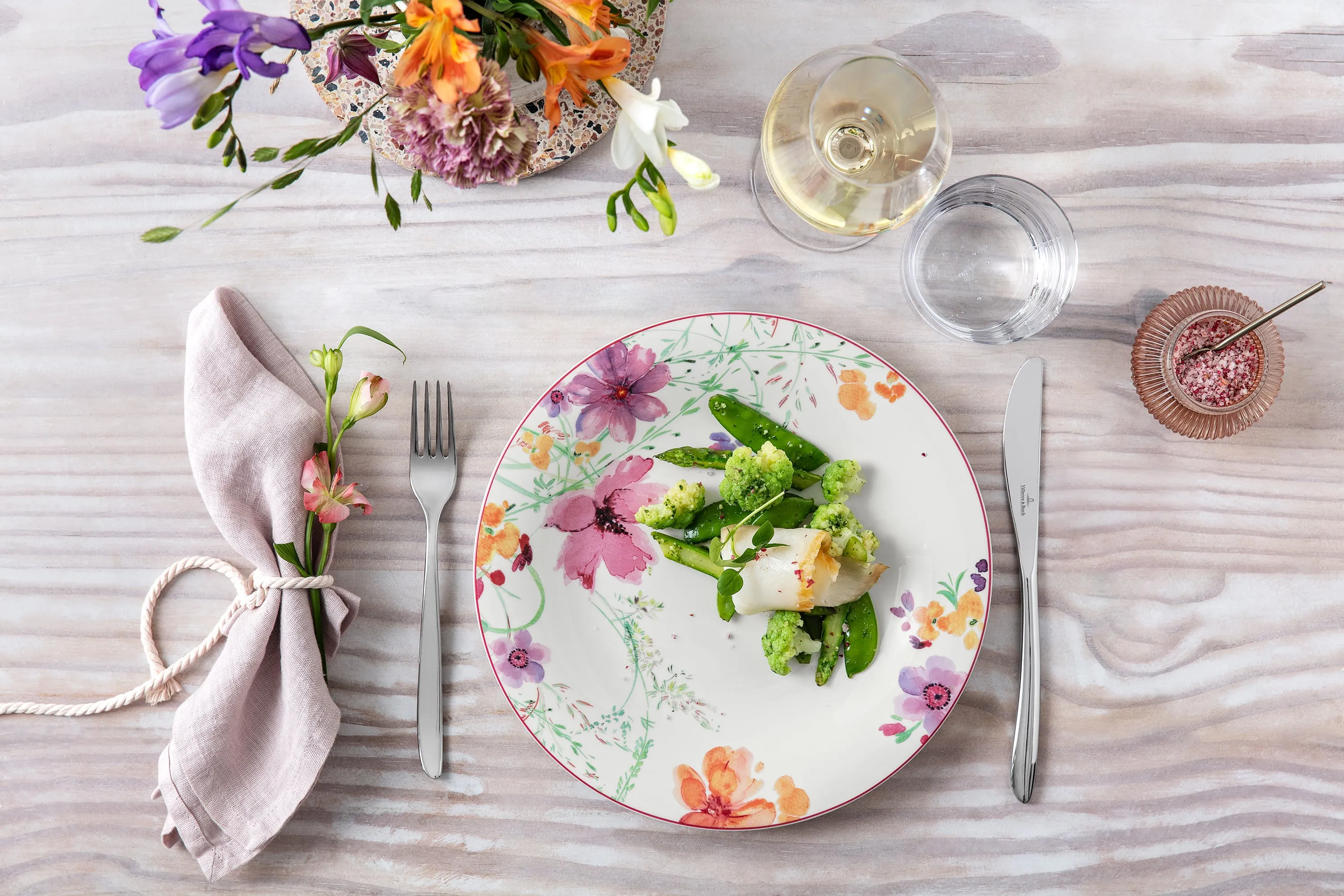 Elegante tavolo da pranzo con un piatto Mariefleur con motivo floreale, un pasto sano e un bicchiere di vino bianco.