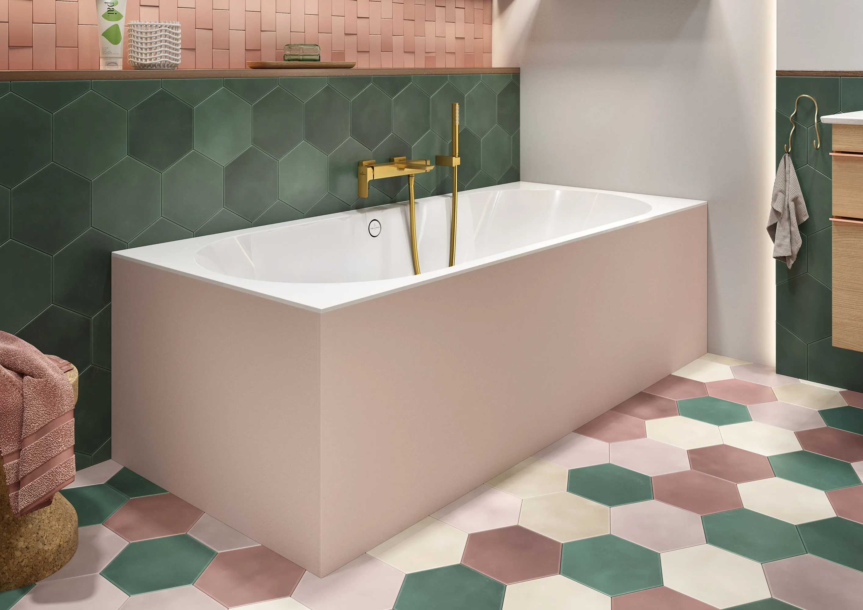 Une salle de bains moderne avec une baignoire blanche revêtue de rose avec une robinetterie dorée devant un mur carrelé en hexagones verts et roses, assortis aux carreaux de sol hexagonaux colorés.