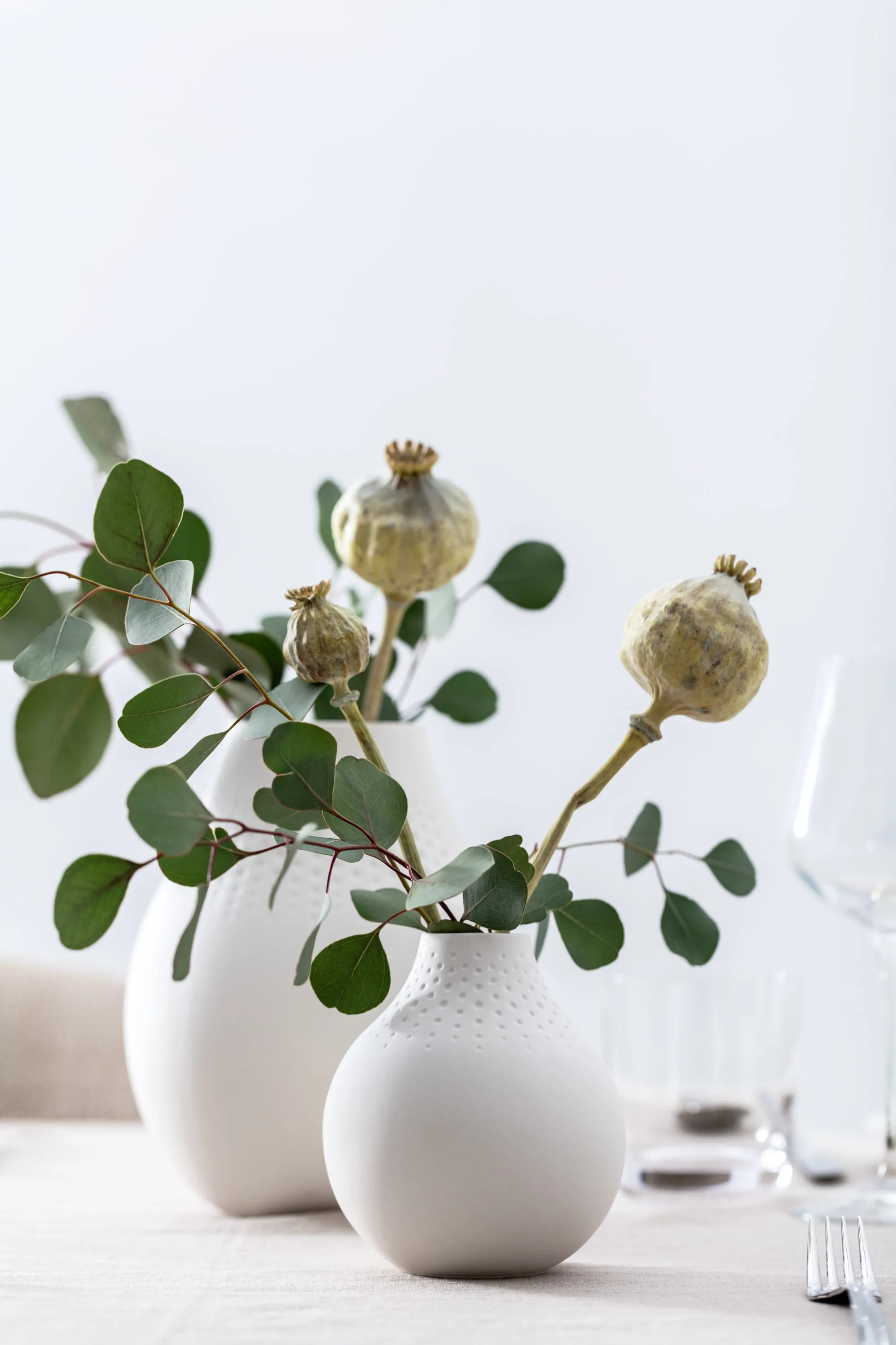 Une décoration de table minimaliste avec une branche d’eucalyptus et des capsules de pavot dans des vases blancs Manufacture.