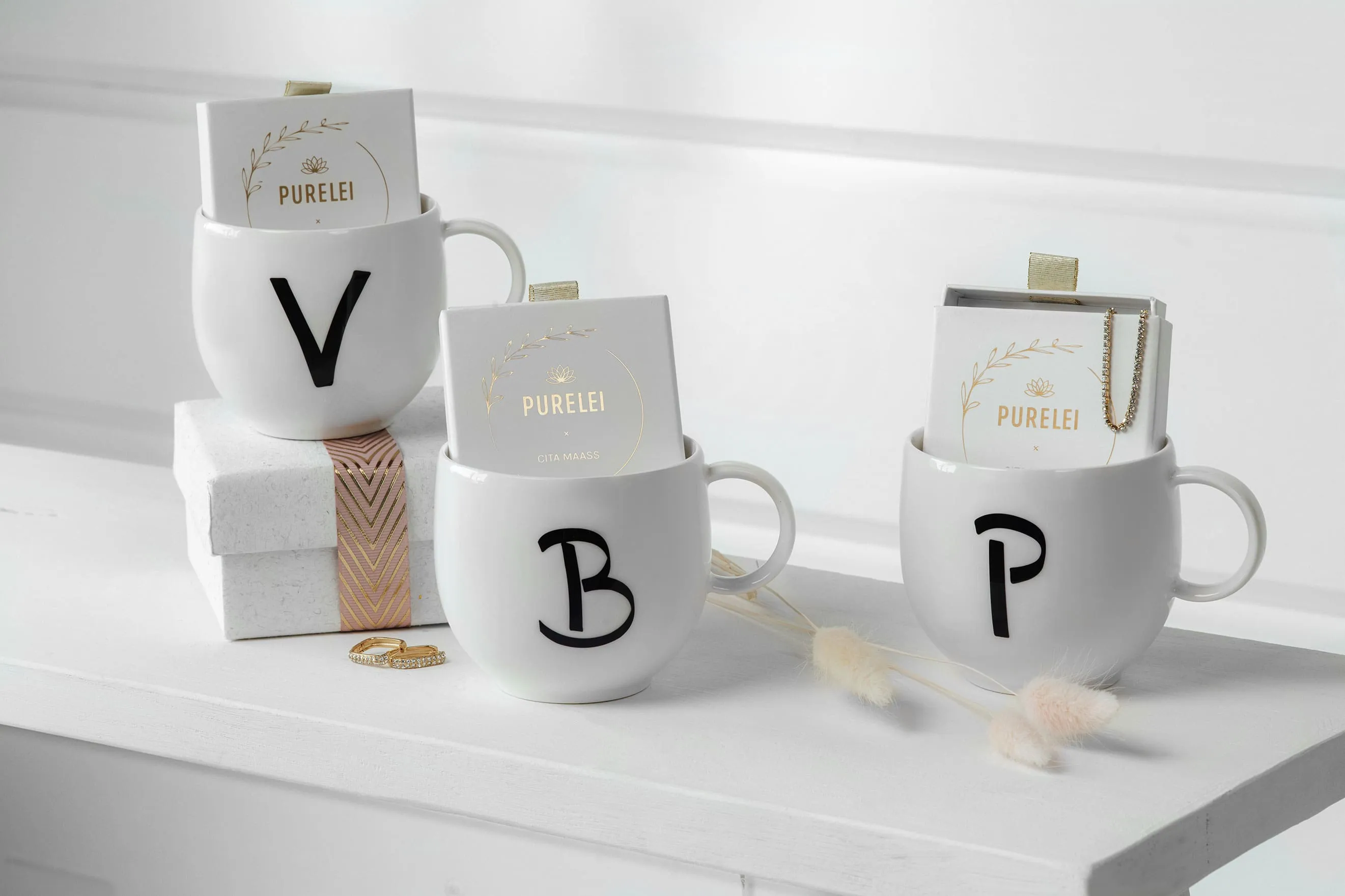 Trois tasses Letters personnalisées portant respectivement les lettres « v », « b » et « p » remplies de bijoux ainsi que des boîtes de produits cosmétiques disposés sur une étagère blanche.