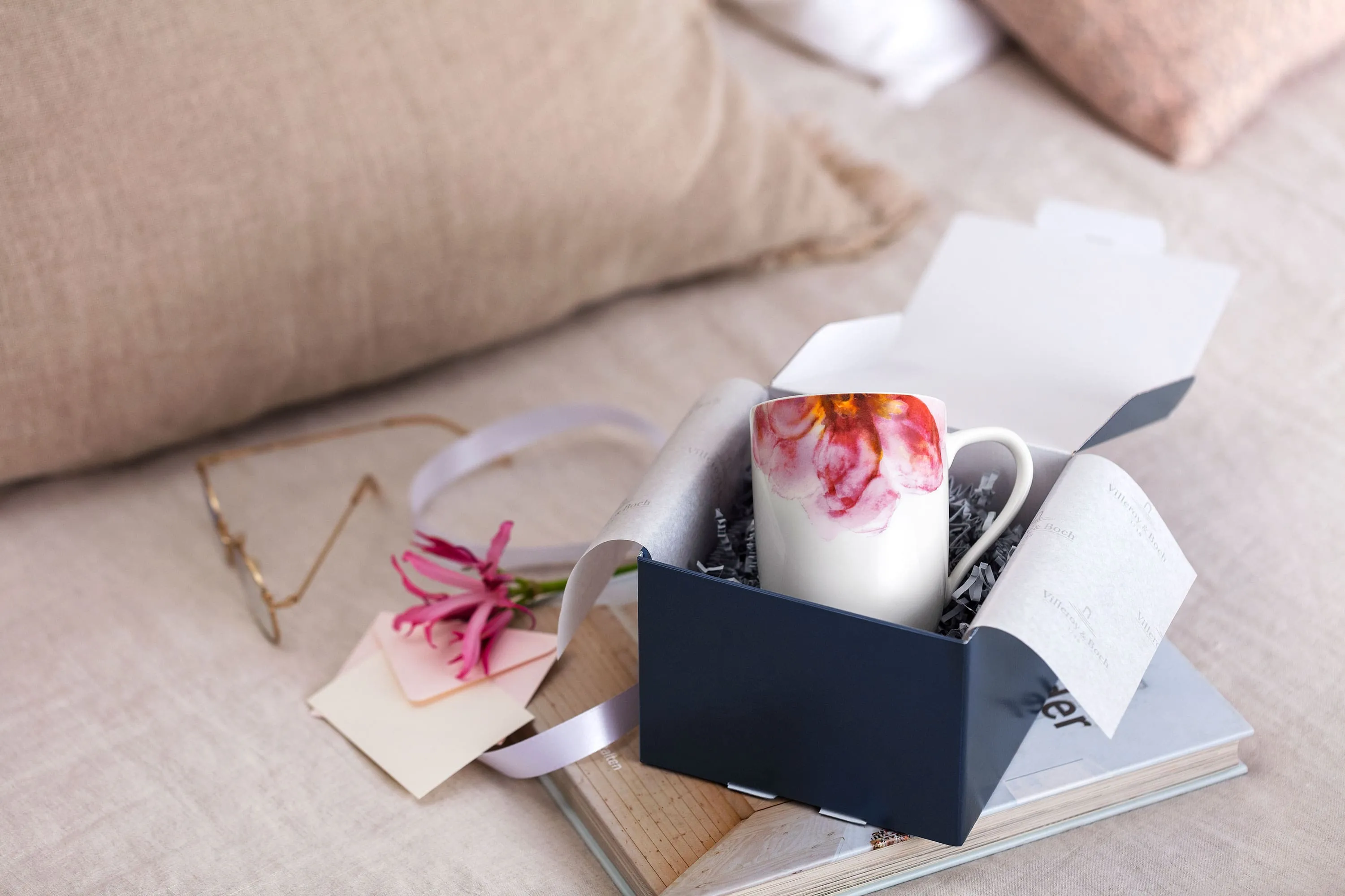 Une boîte cadeau Villeroy & Boch ouverte avec une tasse Rose Garden, des fleurs roses et une note sur un plateau en bois, avec des coussins moelleux en arrière-plan.