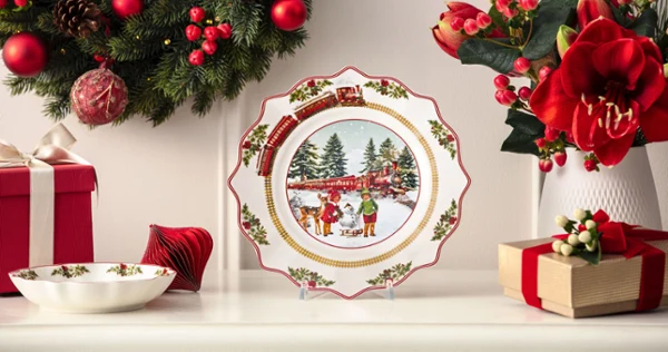 A decorative Christmas plate featuring a winter scene is displayed on a mantelpiece adorned with Christmas garlands, flowers, and wrapped presents.