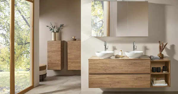 Moderno cuarto de baño con dos lavabos sobre una encimera de madera, armarios de pared, espejos redondos y vistas a los árboles a través de una gran ventana.