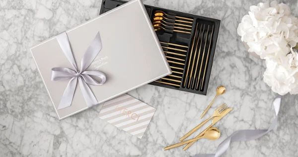 Gift box with gold MetroChic cutlery and a bow Gift box with gold MetroChic cutlery and a bow, a thank you card and white flowers on a marble surface.