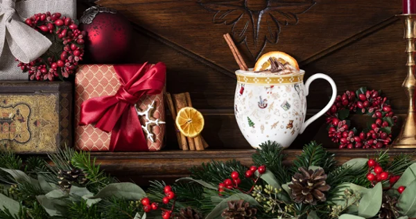 A festive holiday presentation featuring Villeroy & Boch’s Toy’s Delight mug filled with a spiced beverage accompanied by wrapped gifts, ornaments, and greenery on a wooden mantelpiece.