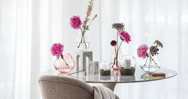 A marble table with various flowers in Rose Garden glass vases and Rose Garden hurricane lamps in front of a window with curtains.