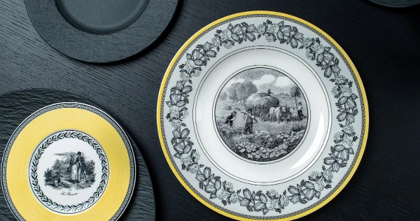 Audun and Manufacture Rock plates on a black table Elegant plates on a dark surface; one large with a rural scene and floral border, another smaller with a yellow rim depicting a couple.