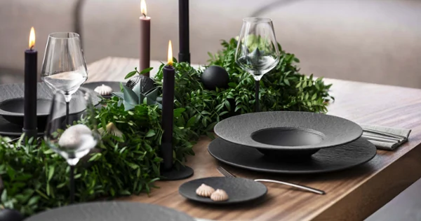Black Manufacture plates on a Christmas table with a modern design A dining table with black Manufacture plates, flatware, wine glasses, and dark candles. Green plants are used as table decorations and create a sophisticated atmosphere.