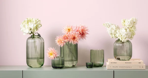 Green Fleur glass vases filled with flowers Green glass vases from Fleur with white and pink flowers arranged on a light green surface in front of a light pink wall.