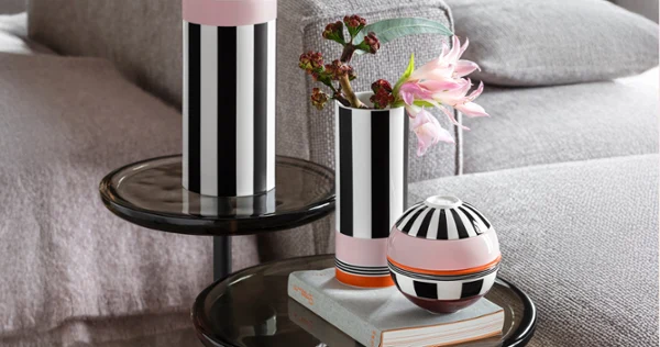 Two black and white striped vases and a round vessel stand on glass tables next to a grey sofa, flowers in a vase