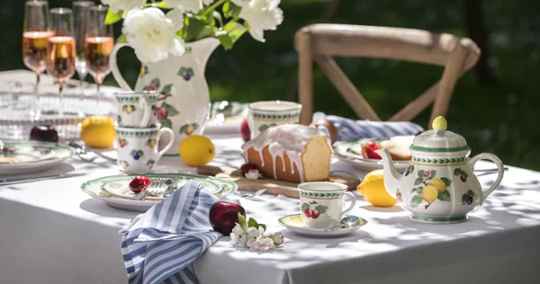 Outdoor table setting with floral French Garden porcelain, a teapot, rosé glasses and a cake. White peonies in a vase and fruit are arranged around the table.