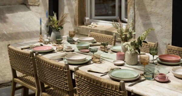 A rustic outdoor dining table setting with woven chairs, like. by Villeroy & Boch pastel-coloured Perlemor plates, glasses and floral centrepieces against the backdrop of a stone wall with a window.