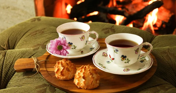 Two floral teacups and saucers on a wooden platter with cookies in front of fireplace