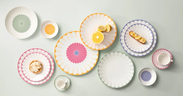 Various colourful, patterned plates, bowls and a cup are arranged on a bright surface. Various colourful, patterned plates, bowls and a cup are arranged on a light-coloured surface, each with a different design. Pastries are placed on some of the plates.