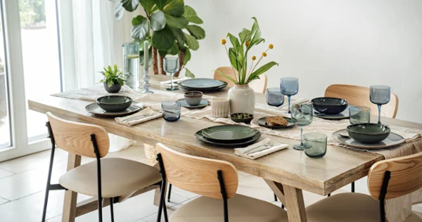 A dining table with green plates, beige napkins, blue glasses and small plants A dining table with green plates, beige napkins, blue glasses and small plants. Light-coloured wooden chairs surround the table. A large plant stands near a window.