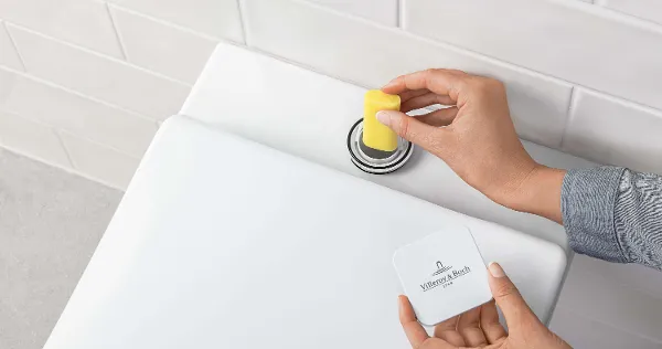 Befüllung der ViFresh Vorrichtung an einer Toilette Eine Person wirft einen gelben WC-Stein in den Spülkasten einer Toilette. Im Hintergrund sind weiß geflieste Wände zu sehen.