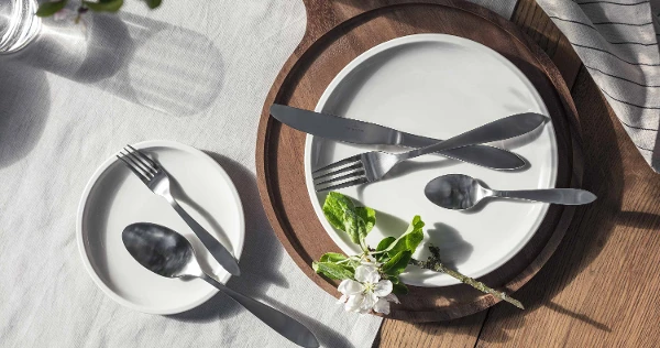 Elegant table decor with simple white plates and Arthur brushed stainless steel flatware, accentuated by natural light and a sprig of flowers.