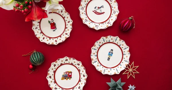 Four decorative Christmas plates with festive motifs on a red tablecloth, surrounded by ornaments and flowers.