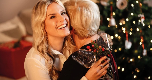 Two women embrace in front of a decorated Christmas tree. The woman looking into the camera is holding a Toy’s Delight wine glass.