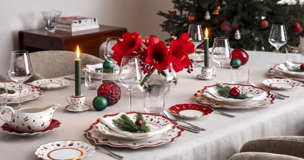 A dining table set for a festive meal A dining table set for a festive meal with red flowers, candles, and festive decorations next to a decorated Christmas tree.