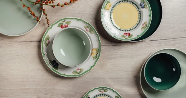 An overhead view of mixed dinnerware, featuring solid green bowls and plates alongside white plates with a colorful fruit border, arranged on a light wooden surface
