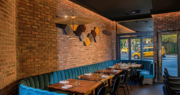 Warm, modern restaurant interior with exposed brick, teal banquettes, wood tables, pendant lighting, geometric art, and a yellow taxi outside.
