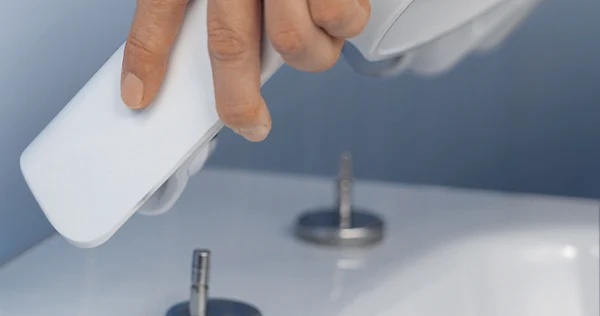 QucikRelease-Funktion für einen WC-Deckel Nahaufnahme einer Hand, die vor einem blauen Hintergrund eine Bidetdüse über einer weißen Badezimmerarmatur mit zwei Bedienelementen hält.