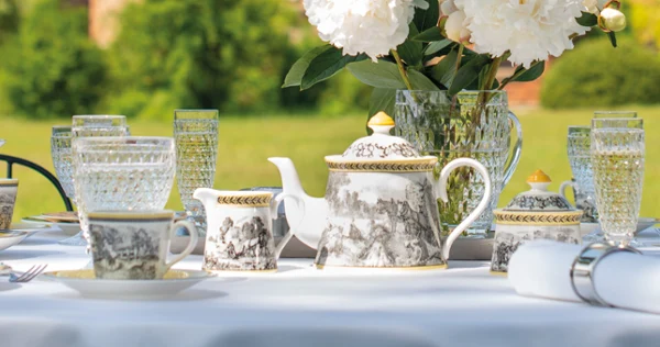 Outdoor tea setting with Audun crockery A table set for outdoor tea with ornate teacups and an Audun teapot, crystal glasses and white peonies in a vase. A blurred historic building is visible in the background.