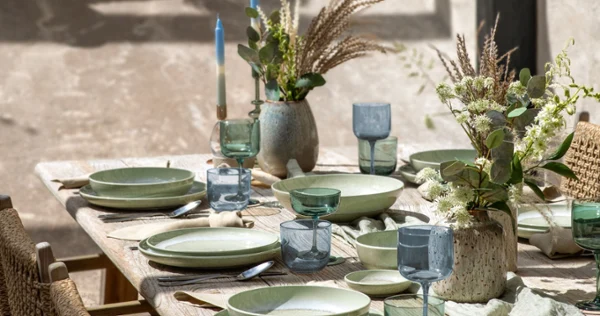 Perlemor Alga Outdoor Dining Table Setting Rustic outdoor dining table set with Perlemor Alga green porcelain dinnerware, blue drinking glasses, woven chairs, and floral centerpieces in a sunlit courtyard.