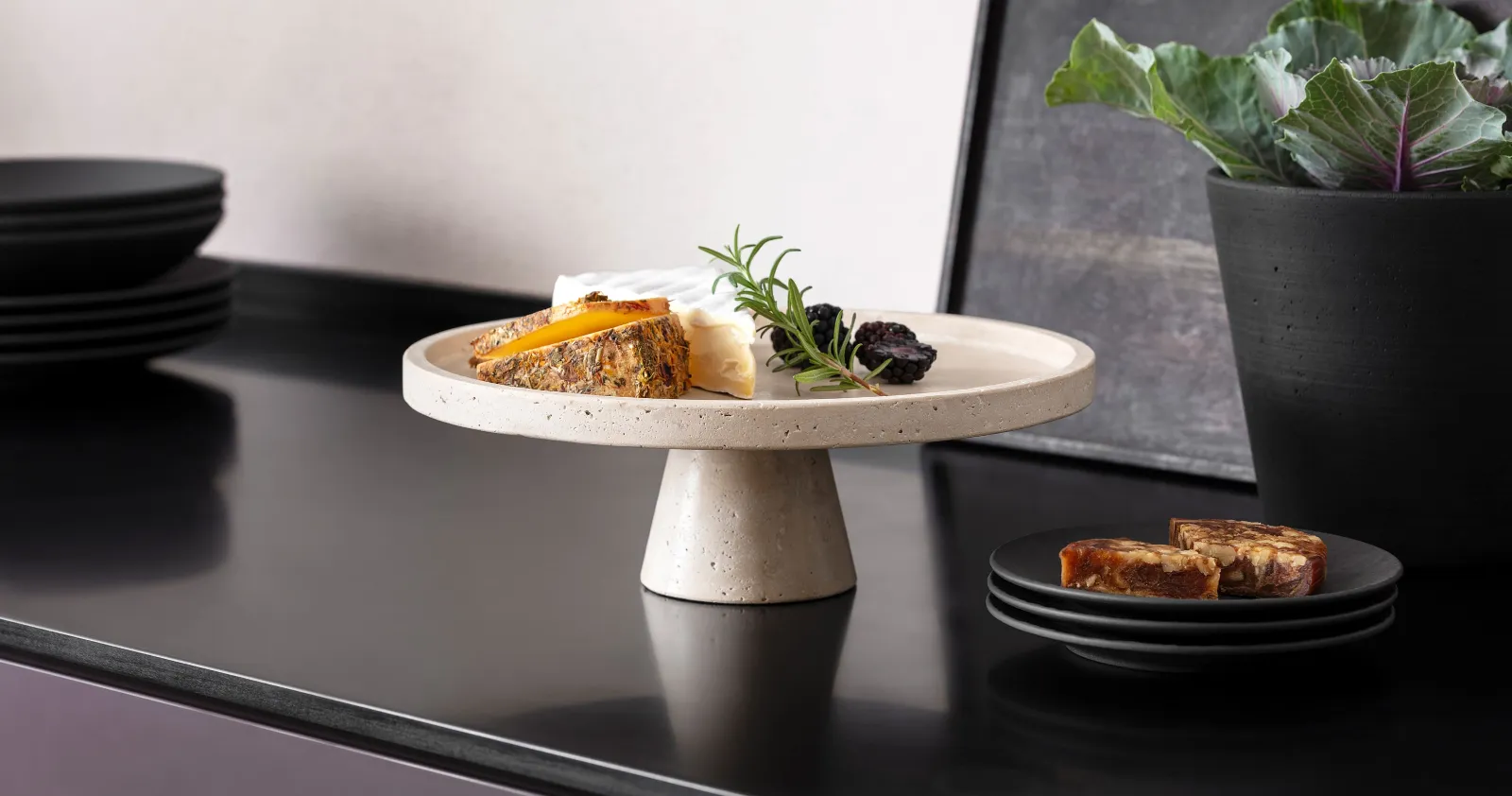 Perfekte Servierschale aus Travertin für Antipasti oder Käse. Käsesortiment auf einer Travertin Servierschale mit Fuß und dunkle Früchten, neben schwarzem Manufacture Geschirr von Villeroy & Boch auf einer dunklen Arbeitsplatte.