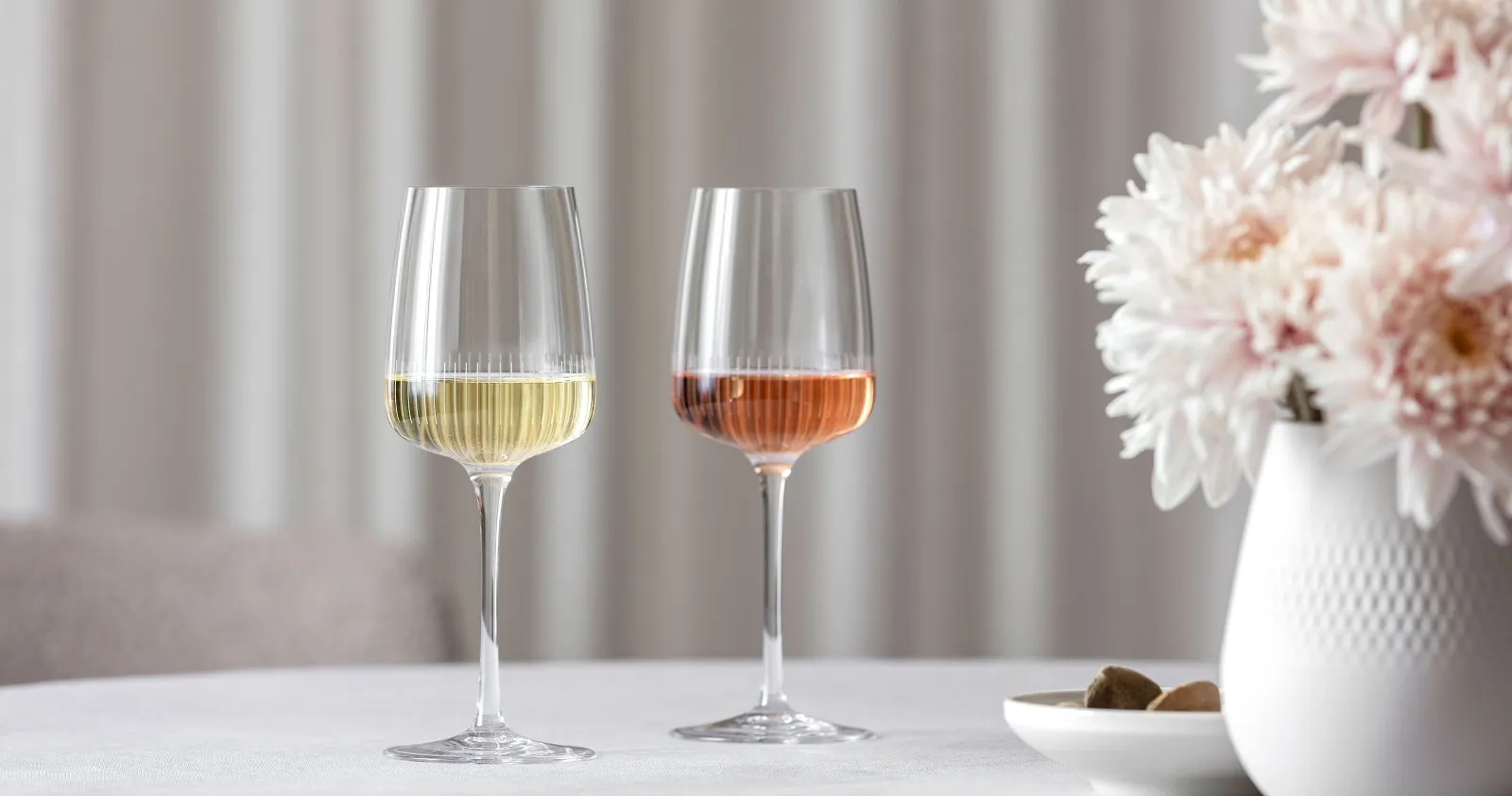 Deux verres à vin Afina de Villeroy & Boch, l'un rempli de vin blanc et l'autre de rosé, sont posés sur une table à côté d'un vase avec des fleurs roses.