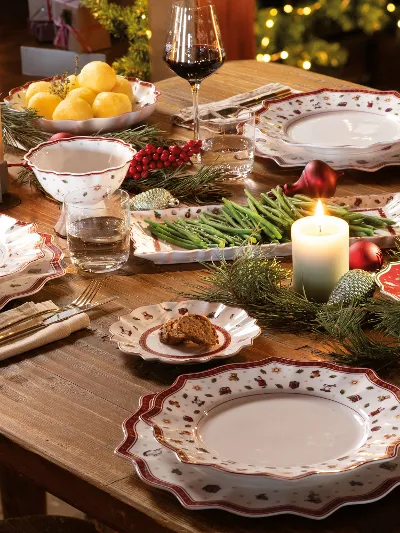 Ein festlicher Esstisch ist mit gemusterten Tellern, Gläsern und Besteck von Villeroy & Boch Toy's Delight gedeckt, die brennende Kerzen und Weihnachtsdekorationen runden das Ambiente ab.