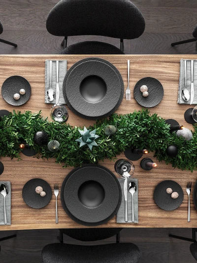 A top view of a dining table set for Christmas with black plates from the Manufacture range by Villeroy & Boch and complemented by Piedmont cutlery. It is also decorated with a centerpiece of green foliage and candles on a wooden surface.