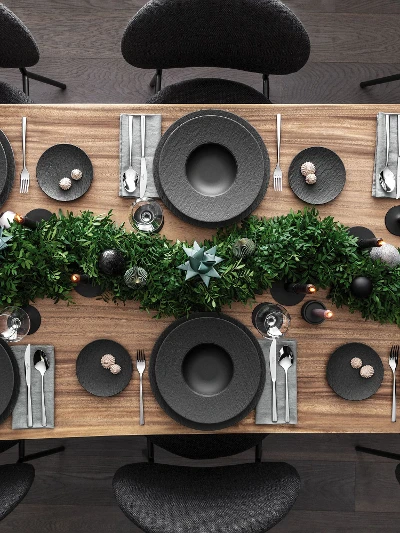 A top view of a dining table set for Christmas with black plates from the Manufacture range by Villeroy & Boch and complemented by Piedmont cutlery. It is also decorated with a centerpiece of green foliage and candles on a wooden surface.