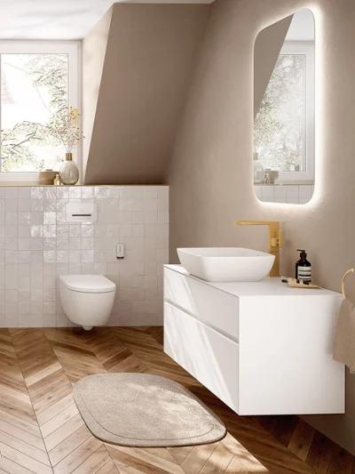 Et moderne beige baderom med et hvitt ViClean I200-toalett med dusjfunksjon, en toppmontert Artis-servant på et Collaro servantskap, en gyllen kran og et opplyst speil over et fiskebensmønstret tregulv.