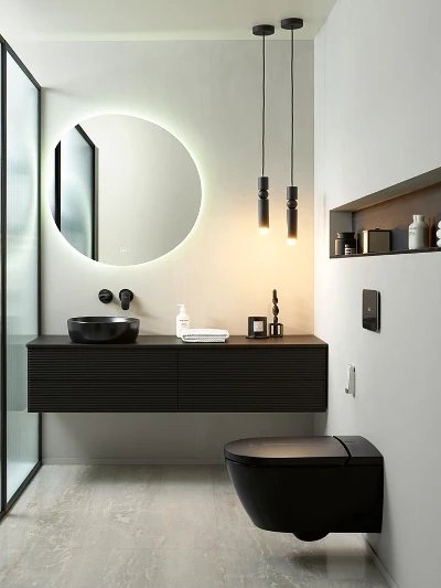 WC e lavabo Antao neri di Villeroy & Boch Bagno moderno con specchio rotondo retroilluminato, mobile lavabo sospeso nero con lavabo da appoggio, WC Antao nero di Villeroy & Boch e arredo minimalista. Due luci a sospensione pendono dal soffitto. Divisorio in vetro a sinistra.