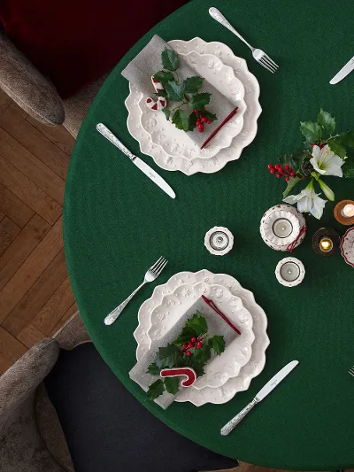 Ein runder Tisch für vier Personen mit weißen Tellern von Villeroy & Boch Toy’s Delight Royal Classic, einer grünen Tischdecke und weihnachtlichen Dekorationen, darunter Stechpalmenblätter, Beeren und Zuckerstangen, von oben betrachtet.