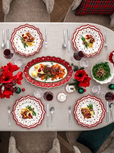 A festive dining table for eight people with red and white patterned plates, red flowers, glasses, flatware, and various dishes, including pasta and salad, on a white tablecloth.