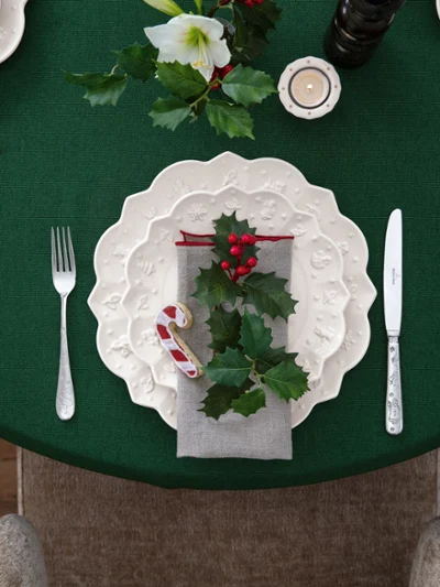 A Christmas table setting with a green tablecloth and white plates from the Villeroy & Boch Toy's Delight Royal Classic collection A Christmas table setting with a green tablecloth, white plates decorated with holly from the Villeroy & Boch Toy's Delight Royal Classic collection, a candy cane, a sprig of holly on the napkin and silver cutlery.