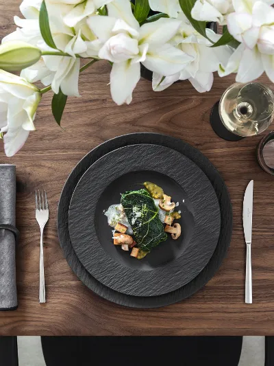 Manufacture von Villeroy & Boch für edle Anlässe Elegantes Tischgedeck mit schwarzen Manufacture Tellern, Besteck, Feigen, einem Gericht mit Gemüse und Garnelen, grauen Servietten, weißen Blumen, einem Weinglas und geschnittenem Brot.