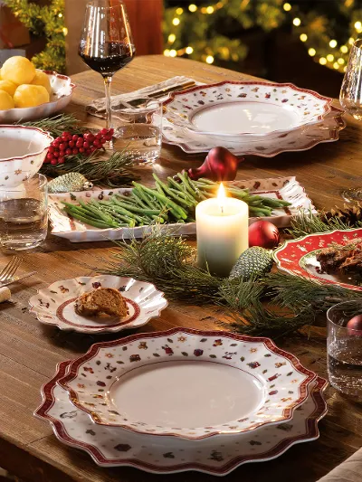 Ein festlicher Esstisch ist mit gemusterten Tellern, Gläsern und Besteck von Villeroy & Boch Toy's Delight gedeckt, die brennende Kerzen und Weihnachtsdekorationen runden das Ambiente ab.