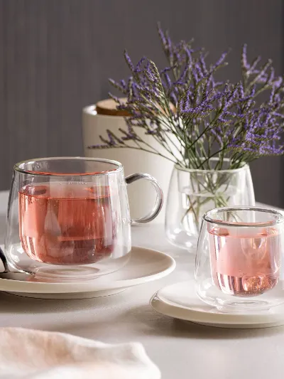Artesano glass cups filled with pink tea, placed on saucers next to a spoon on a table. Artesano glass cups from Villeroy & Boch filled with pink tea, placed on saucers next to a spoon on a table that is decorated with a small vase of purple flowers.