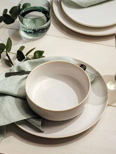 A white Crafted Bol on a Crafted plate covered with mint green napkins and eucalyptus.